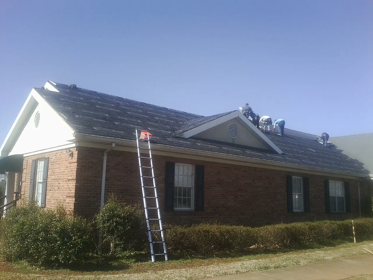 Crew tearing shingles off a brick ranch roof with a ladder set — Emergency Roof Repair work by our crew in Sagamore Beach