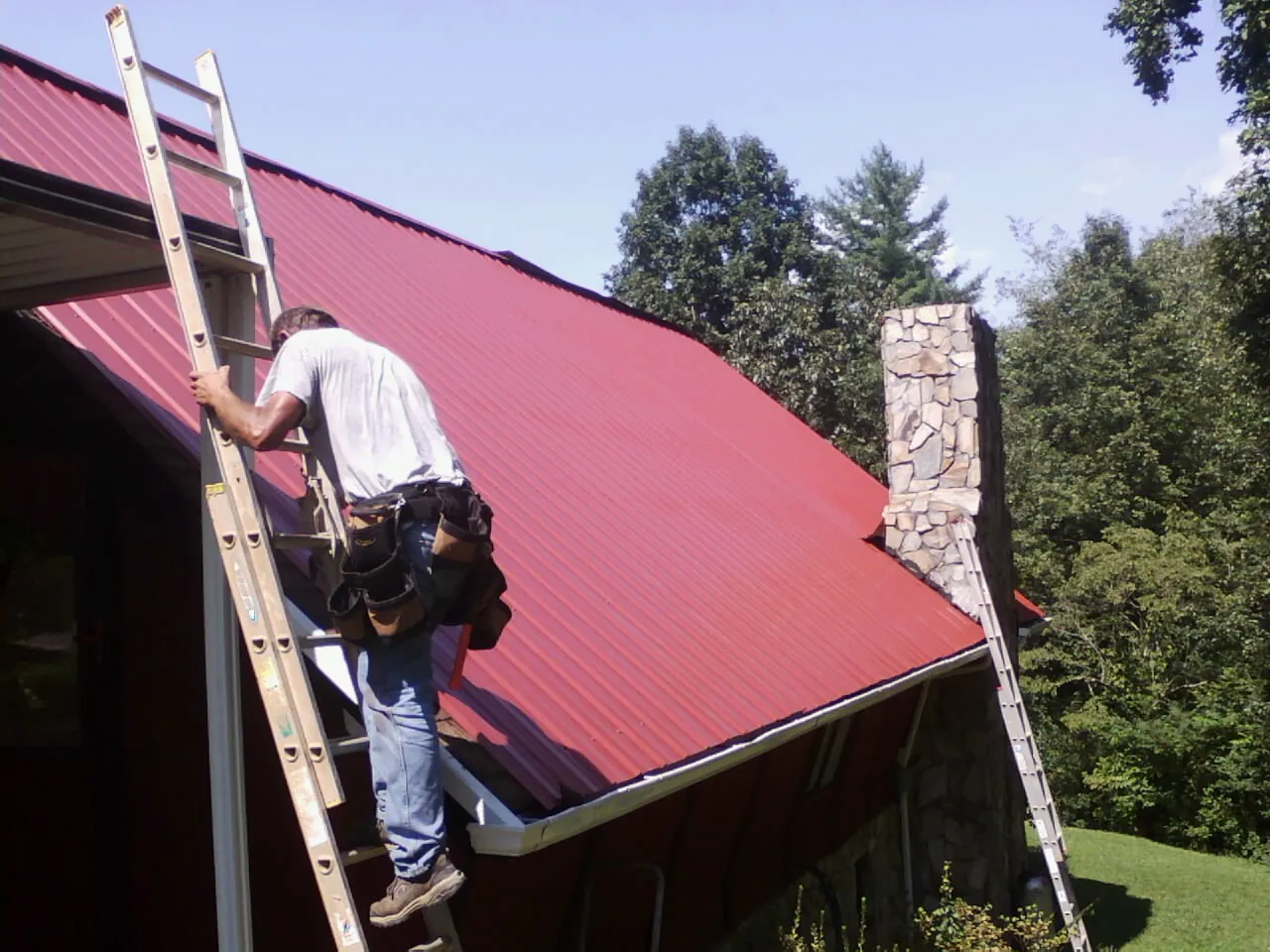 Roofer on a ladder beside a red metal roof and stone chimney — Soffit & Fascia Repair by Roofing Brothers in Sagamore Beach